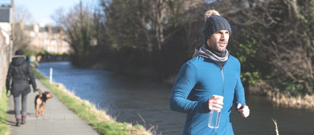 A man wearing a fitted hooded top, leggings and a woolly hat jogs on a footpath along a canal in the city of Edinburgh, Scotland, United Kingdom, with the sun on his face while he carries water