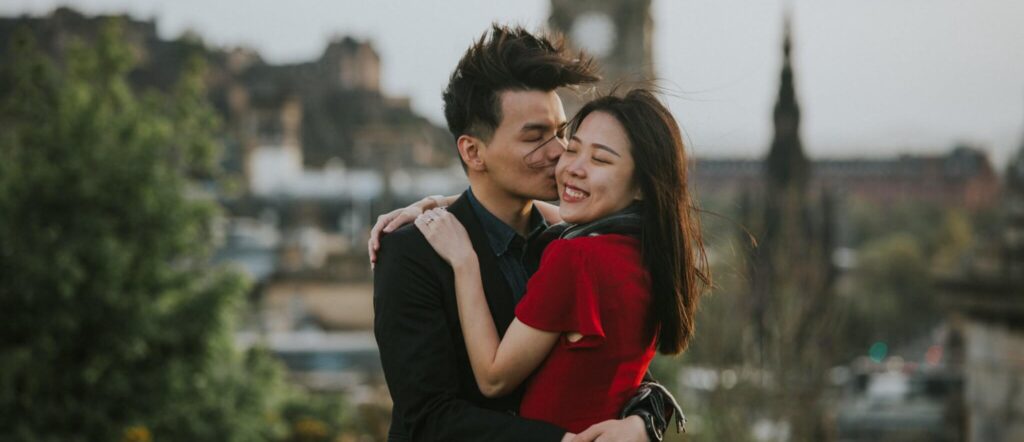 Happy couple celebrating engagement on top of Calton Hill Edinburgh with a blurred out view of the city in the background